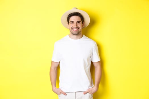 Young handsome guy tourist looking happy, wearing straw hat for travelling, standing against yellow background. Copy space