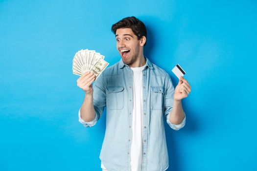 Amazed and happy man holding credit card, looking at money satisfied, standing over blue background.