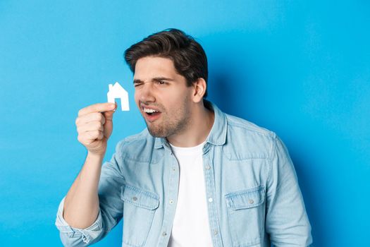 Real estate concept. Man looking at tiny house maket with confused face, standing puzzled against blue background.