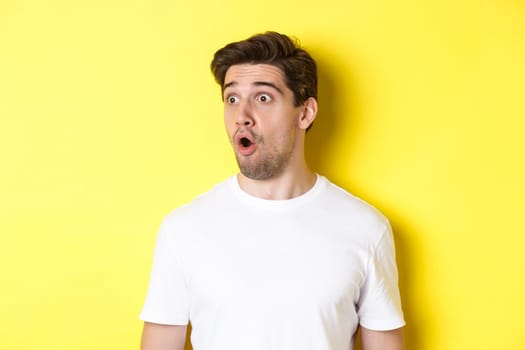 Close-up of impressed man looking left, gasping amazed, standing in white t-shirt against yellow background.