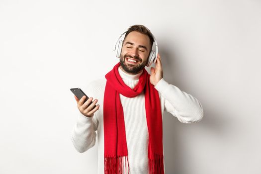 Winter holidays and technology concept. Satisfied man listening music in headphones with closed eyes, smiling with pleasure, holding smartphone, white background.