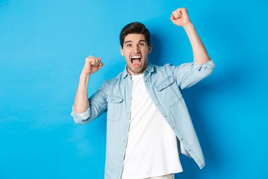 Cheerful guy making fist pumps and rooting for someone, shouting for joy, triumphing over win, standing against blue background.