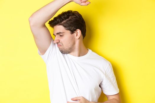 Man in sweat smelling his armpit, standing in white t-shirt and grimacing from stinky clothes.