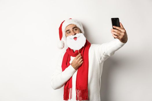 Christmas party and celebration concept. Young man taking selfie with funny white beard Santa mask and hat, posing for photo on mobile phone, studio background.