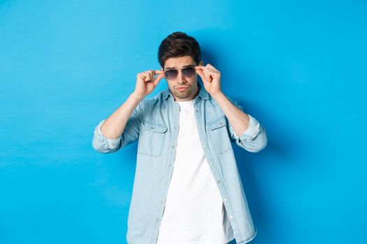 Confident macho man put on sunglasses, looking cool and sassy, standing over blue background.