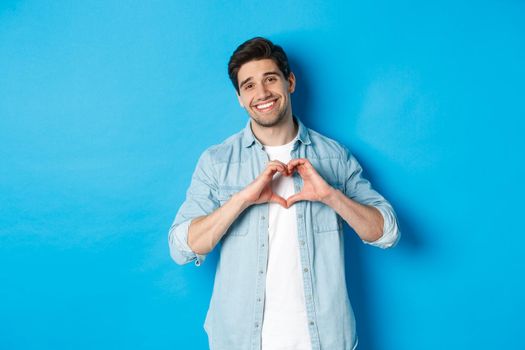 Handsome man smiling, showing heart gesture and looking at camera, saying I love you, standing against blue background.