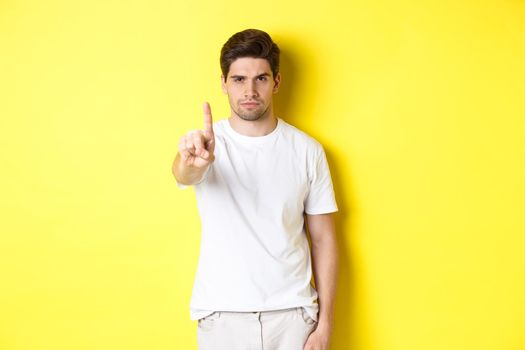 Confident man saying no, decline and prohibit something, showing one finger and frowning, standing over yellow background.