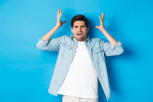 Confused and bothered man holding hands near head, looking frustrated, standing upset against blue background.