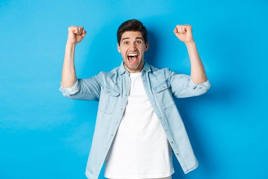 Happy attractive man triumphing, screaming yes and raising hands up to celebrate win, achieve goal, standing against blue background.