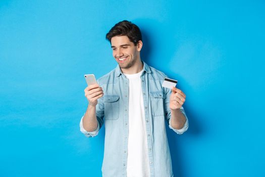 Young man shopping online with mobile application, holding smartphone and credit card, standing over blue background.