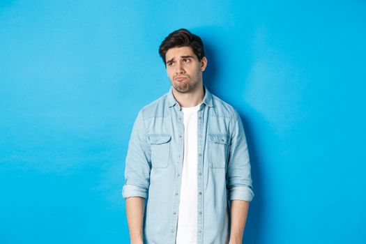 Image of reluctant and sad guy in casual outfit looking left, frowning and feeling upset, standing against blue background.