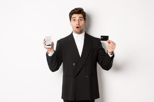 Image of handsome businessman in black suit, looking amazed and showing credit card with mobile phone screen, standing against white background.