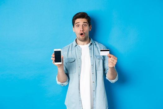 Amazed young man showing mobile cell phone screen and credit card, shop online, standing against blue background.