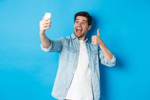 Happy man taking selfie and showing thumb up in approval on blue background, holding mobile phone.