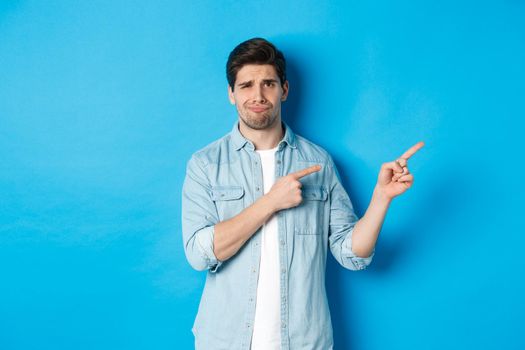 Portrait of skeptical adult guy pointing fingers right and smirking, exress disappointment and doubt, standing against blue background.