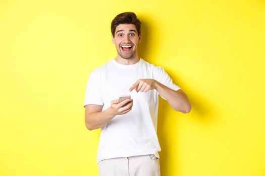 Happy man pointing at smartphone to show promo, check out internet offer, standing over yellow background. Copy space