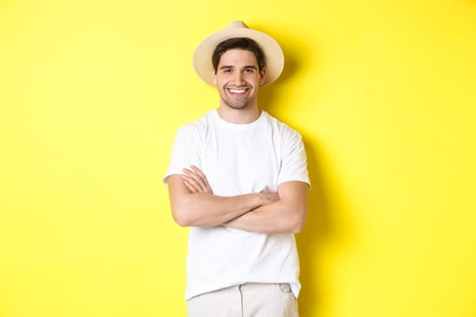 Tourism. Handsome young man looking happy, wearing straw hat for travelling, cross hands on chest and smiling, standing over yellow background.
