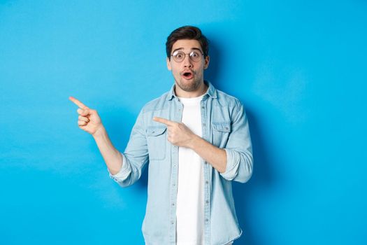 Surprised man in glasses pointing fingers left at copy space, asking question about product, looking intrigued, standing over blue background.