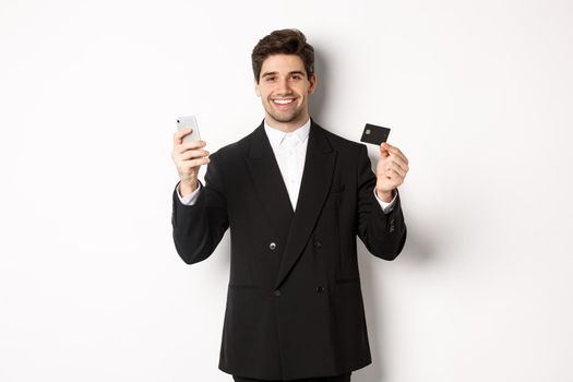 Handsome businessman in black suit smiling, showing credit card and money, standing against white background.