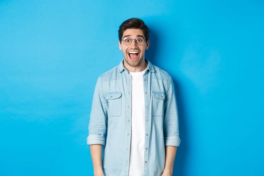 Attractive happy man in glasses looking surprised, checking out advertisement, standing over blue background.