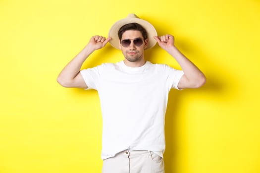 Confident young male tourist ready for vacation, wearing straw hat and sunglasses, standing against yellow background.