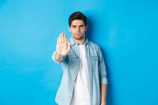 Serious man frowning and saying no, extending hand to shop stop sign, prohibit action, standing against blue background.