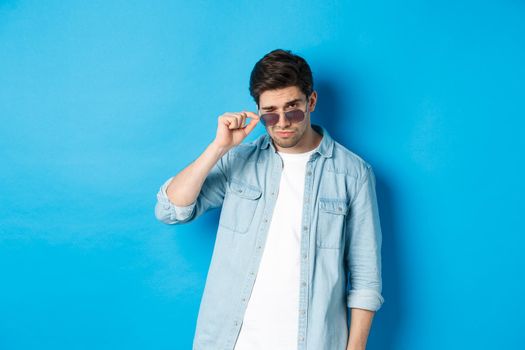 Confident macho man put on sunglasses, looking cool and sassy, standing over blue background.