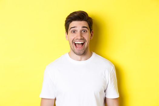 Close-up of surprised handsome guy reacting to great news, standing over yellow background in white t-shirt.