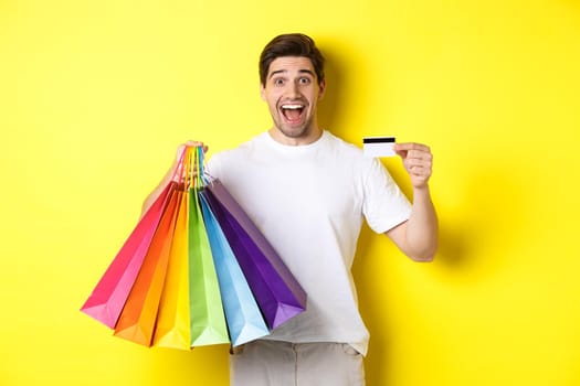 Happy attractive man holding shopping bags, showing credit card, standing over yellow background.