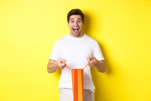 Surprised guy open shopping bag with fist, looking excited and happy at camera, standing against yellow background.