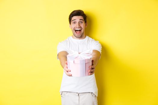 Concept of holidays and celebration. Happy man wishing happy new year, giving you gift, standing over yellow background.