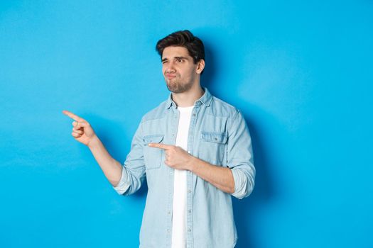 Doubtful adult man pointing fingers left at promotion and looking unsure, grimacing disappointed, standing against blue background.