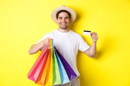 Happy attractive guy showing shopping bags and credit card, concept of banking and easy payment, standing over yellow background.