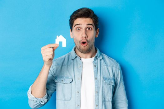 Real estate concept. Surprised guy showing small paper house model, looking amazed, standing against blue background.