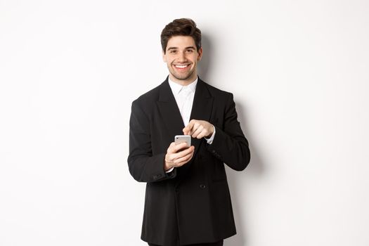 Image of handsome businessman in suit showing you app on smartphone, pointing finger at mobile screen and smiling, standing over white background.