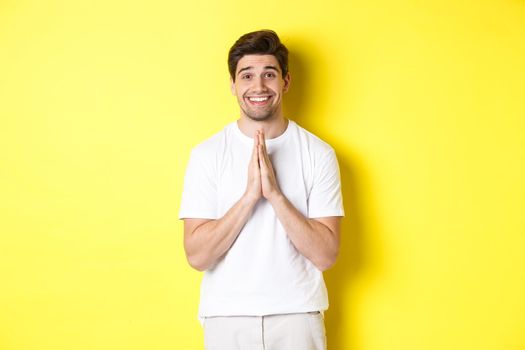 Hopeful man asking for help, begging favour, need something and smiling, standing over yellow background.