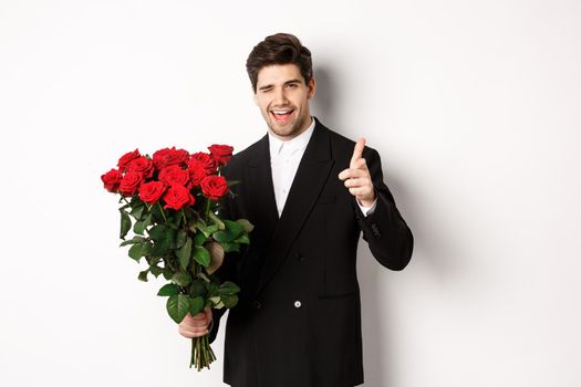 Image of handsome romantic guy in black suit, holding bouquet of roses and pointing at camera, congratulating with holiday, standing against white background.