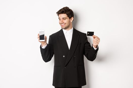 Handsome successful businessman, looking at smartphone screen and showing credit card, standing in black suit against white background.