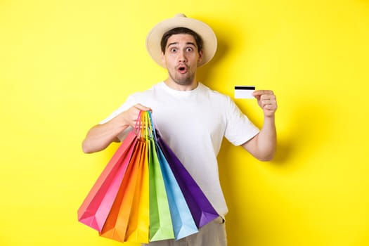 Impressed man showing shopping bags with products and credit card, standing over yellow background.