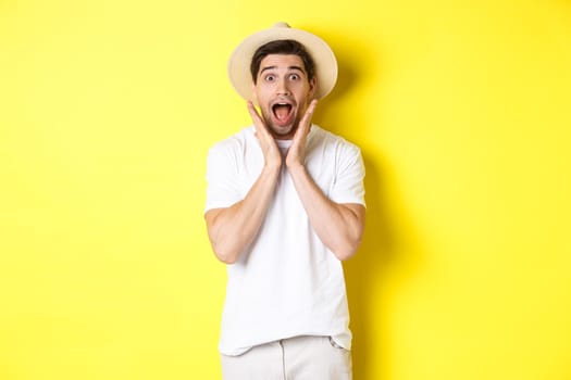 Concept of tourism and summer. Surprised male model in straw hat, looking amazed at special offer, standing against yellow background.
