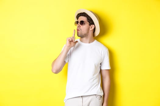 Cool young male tourist blowing at finger gun and looking confident, standing against yellow background. Vacation and lifestyle concept.