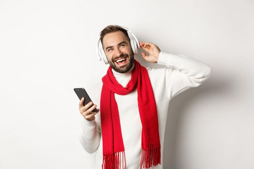 Winter holidays and technology concept. Man enjoying listening music in headphones, looking satisfied, holding smartphone, wearing sweater with scarf, white background.