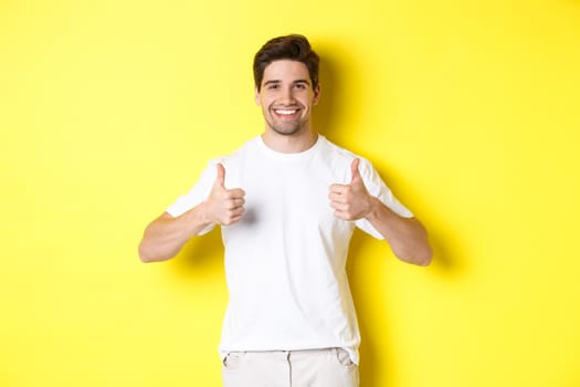 Portrait of happy man showing thumbs-up in approval, like something or agree, standing over yellow background.