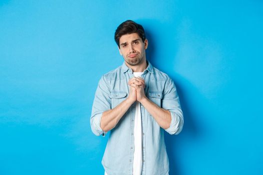 Young guy begging for help, pouting and looking sad while asking for favour, need something, standing against blue background.