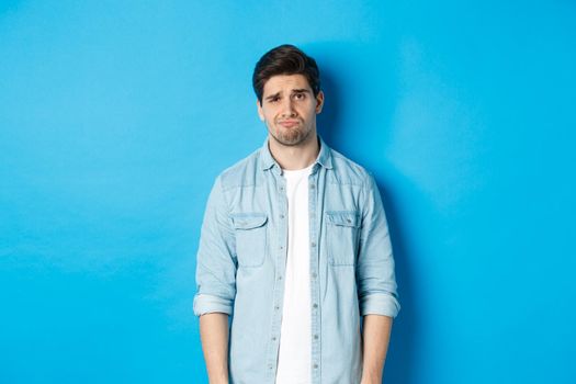 Man looking with doubt at camera, frowning and sulking, feeling uneasy about something, standing against blue background.