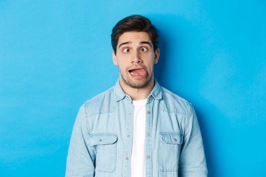 Close-up of young man making funny expressions, showing tongue and looking at camera, standing over blue background. Copy space