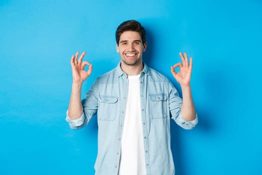 Confident smiling adult man, showing ok signs and looking pleased, like something, standing against blue background.