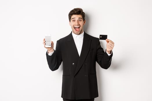 Image of handsome businessman in black suit, looking excited and showing credit card with mobile phone screen, standing against white background.