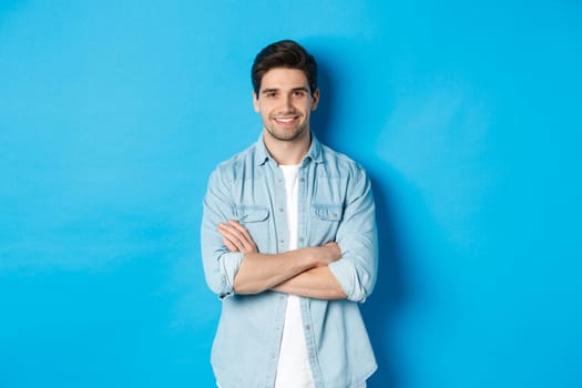 Handsome guy in casual clothes standing with arms crossed and confident smile against blue background.