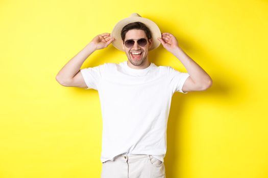 Happy man on vacation, wearing straw hat and sunglasses, smiling while standing against yellow background.